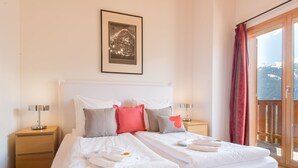 En pænt redt seng med hvide sengetøj og mange puder, et billede i ramme på væggen, og udsigt til en balkon med rødt gardin.