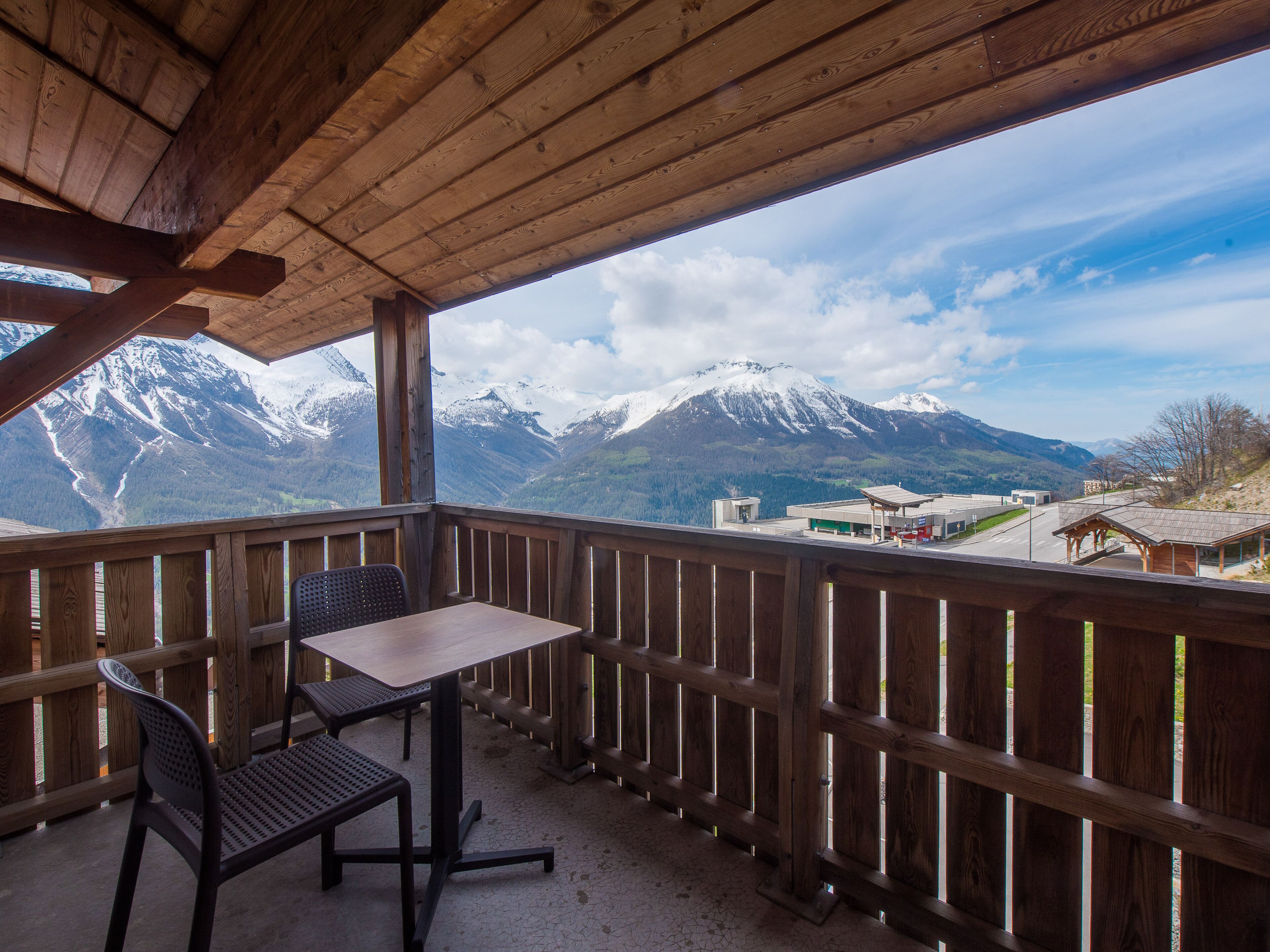 Un balcón de madera con una mesa y sillas, con vistas a montañas nevadas y un edificio pequeño.