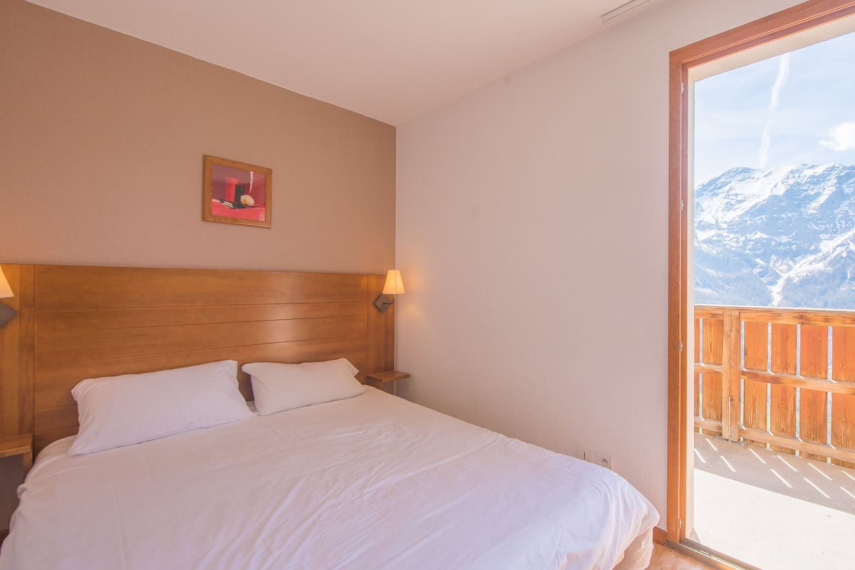 Un dormitorio con cabecera de madera, cama con ropa blanca, mesita de noche con lámpara y una ventana grande con vistas a la montaña.