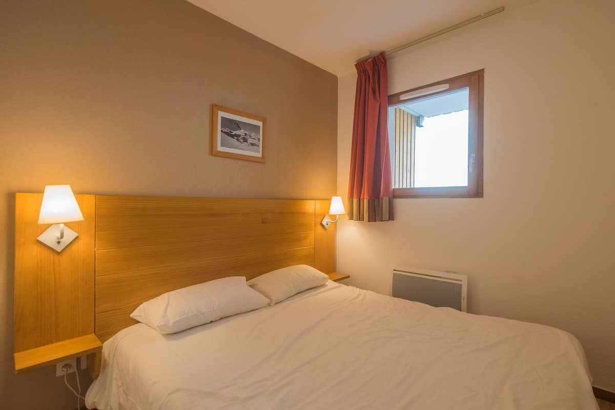 Una habitación de hotel con cabecera de madera, dos lámparas de noche, una cama con ropa blanca, un cuadro en la pared y una ventana con cortina roja.
