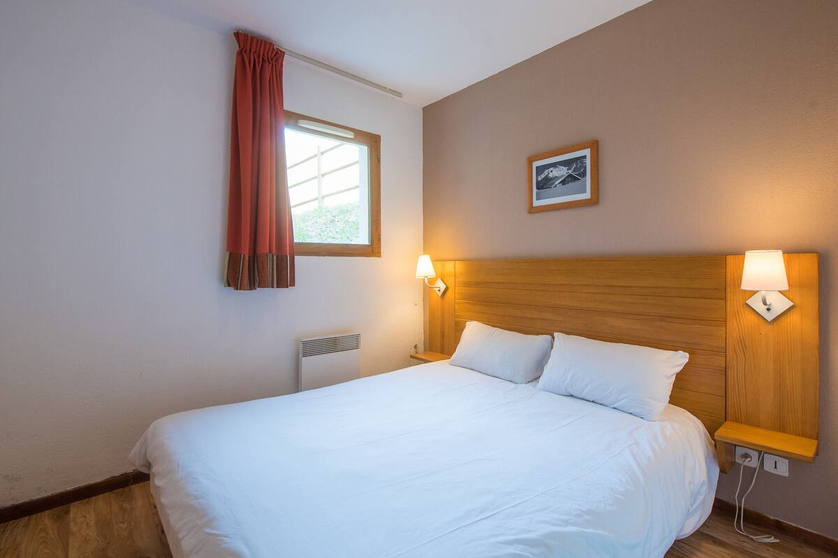 Una habitación de hotel con cabecera de madera, una cama grande con ropa blanca, una ventana con cortina roja y un cuadro colgado en la pared.