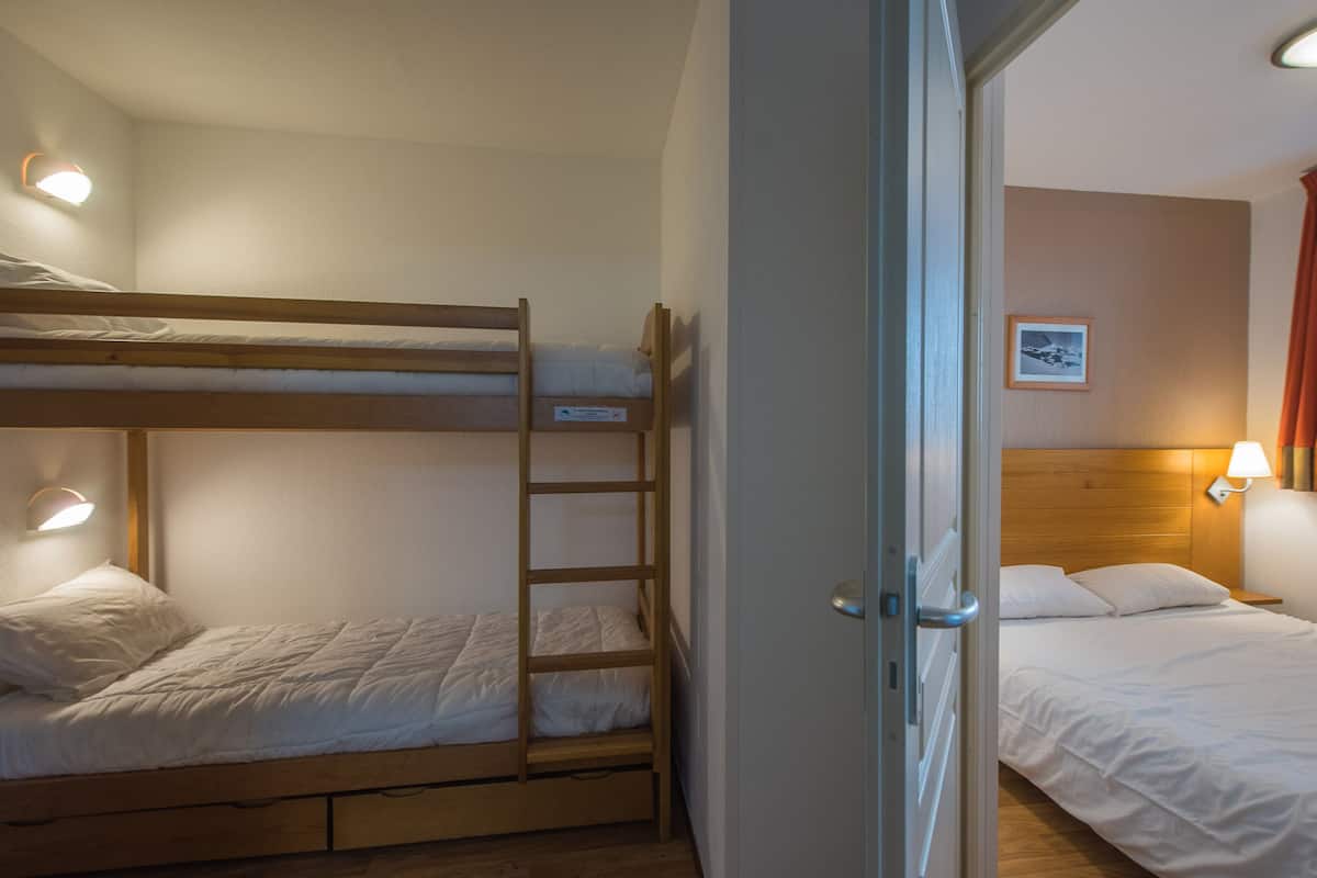 Una habitación con una cama litera, una cama individual, una escalera de madera, una puerta y una lámpara fijada a la pared.