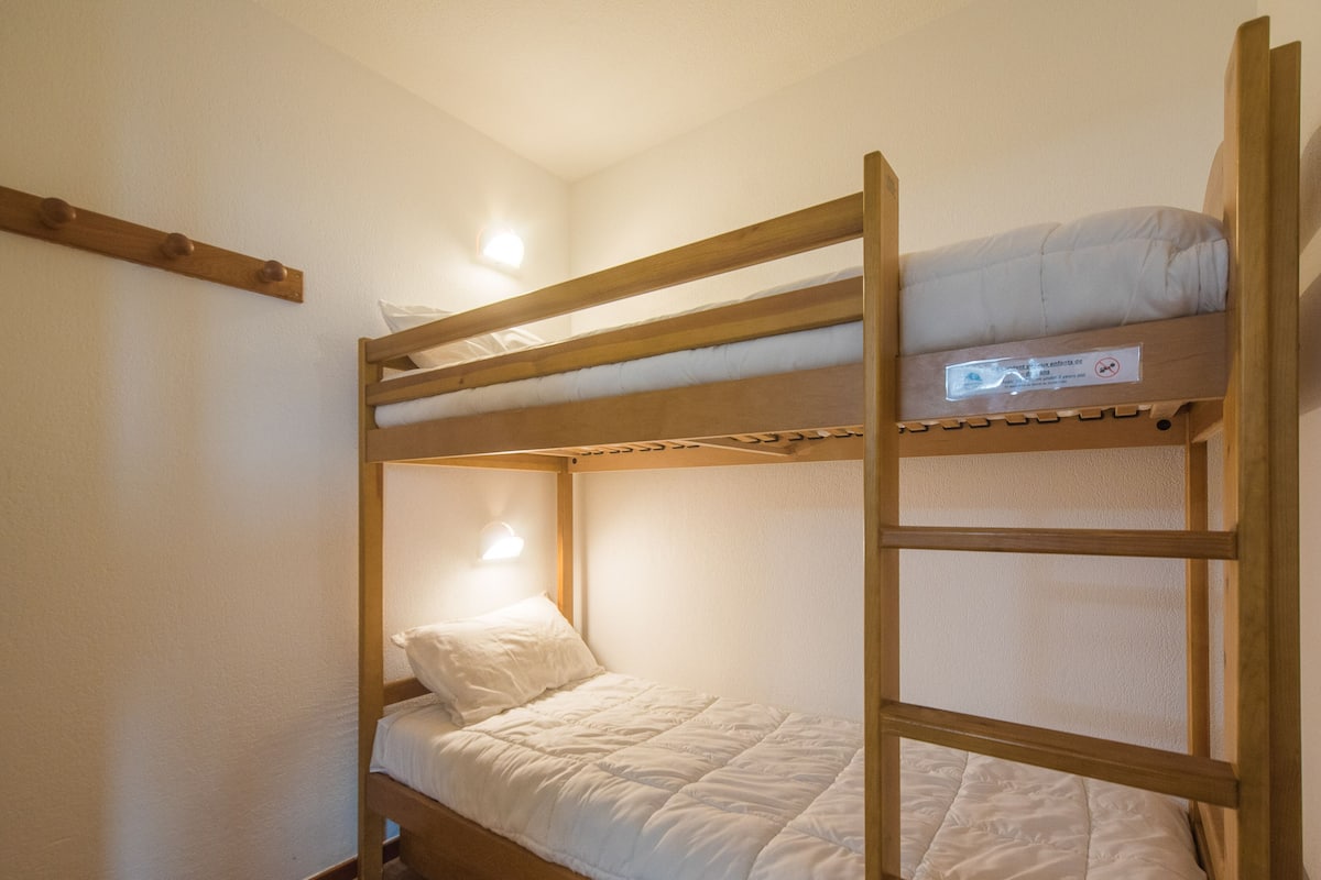 Una cama litera con estructura de madera, ropa de cama blanca y una pequeña lámpara fijada a la pared.