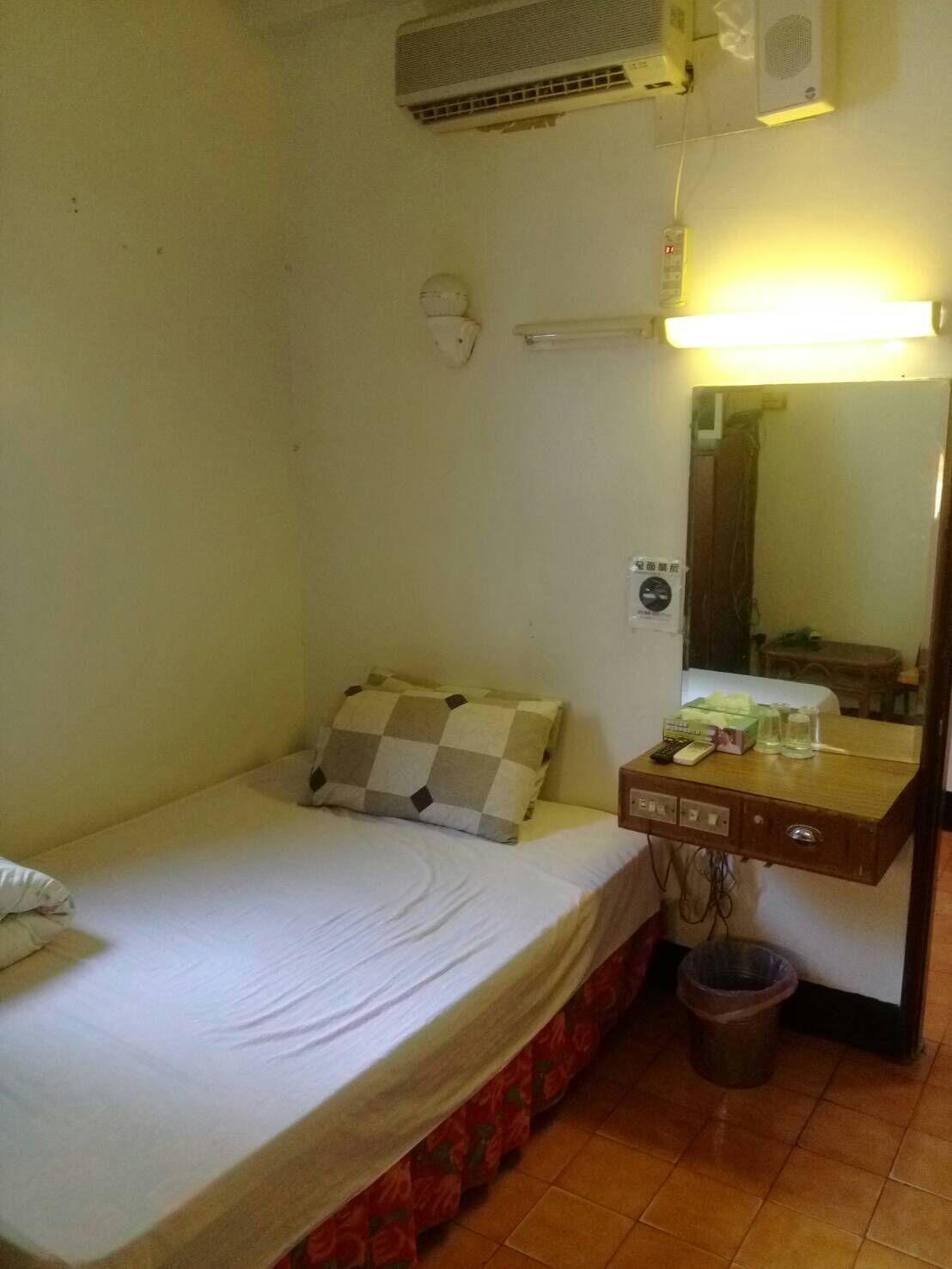 Una habitación pequeña con cama individual, una mesita de noche, un espejo y un cesto de basura.