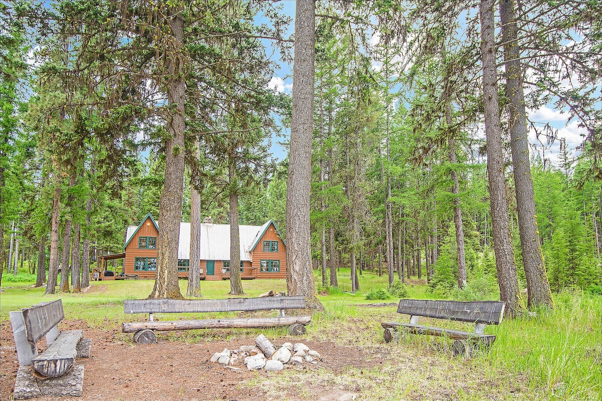 Une cabane entourée de grands arbres, avec deux bancs et un foyer au premier plan.