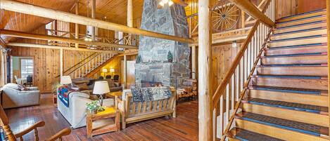 A spacious log cabin interior with a stone fireplace, wooden staircase, and a cozy seating area.