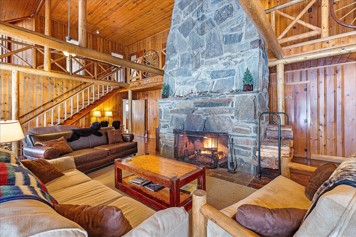 L’intérieur d’une cabane en rondins, avec une cheminée en pierre, un escalier en bois et un coin salon confortable agrémenté de canapés en cuir et d’une table basse.