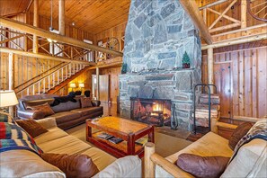 A log cabin interior with a stone fireplace, wooden staircase, and a cozy seating area with leather sofas and a coffee table.