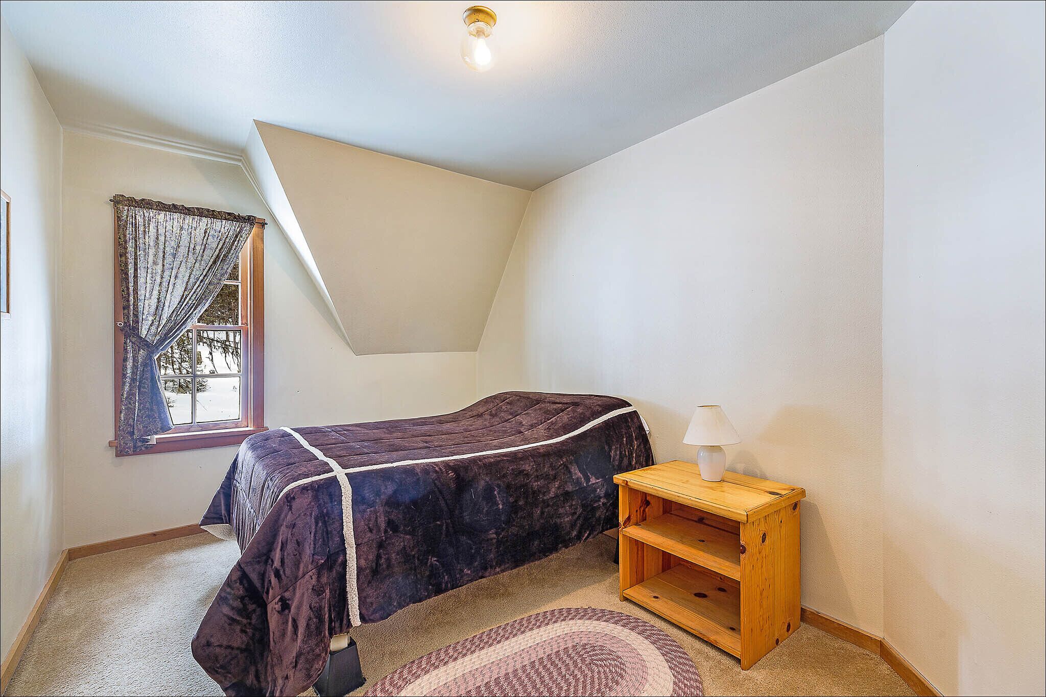 A bedroom with a bed, a nightstand, a lamp, a window with curtains, and a ceiling light.