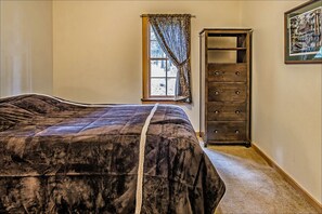 A bedroom with a large bed, a window with curtains, a tall wooden dresser, and a framed picture on the wall.