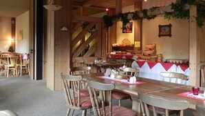 Un espace repas avec des tables et des chaises en bois, un escalier et un coin cuisine avec une nappe rouge.