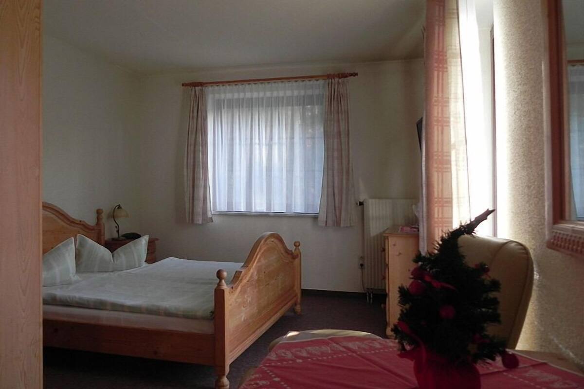 Quarto com cama de madeira, janela com cortinas leves, radiador e uma pequena árvore de Natal.