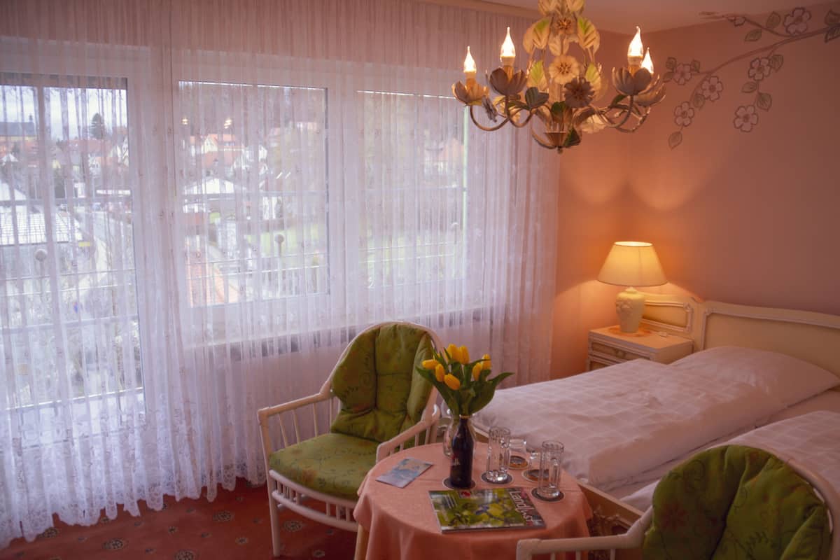 Ein Hotelzimmer mit zwei Einzelbetten, einem grünen Sessel, einem kleinen Tisch mit Blumenvase und einem Fenster mit transparenten Vorhängen.