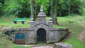 Edificio in pietra con tetto a punta e una porta, immerso nel verde e affiancato da un sentiero.