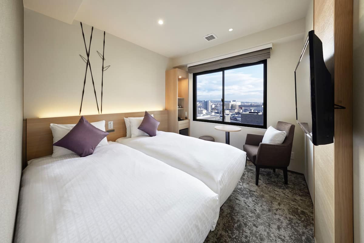 A hotel room with two beds, a small table, a chair, a TV, and a large window with a city view.