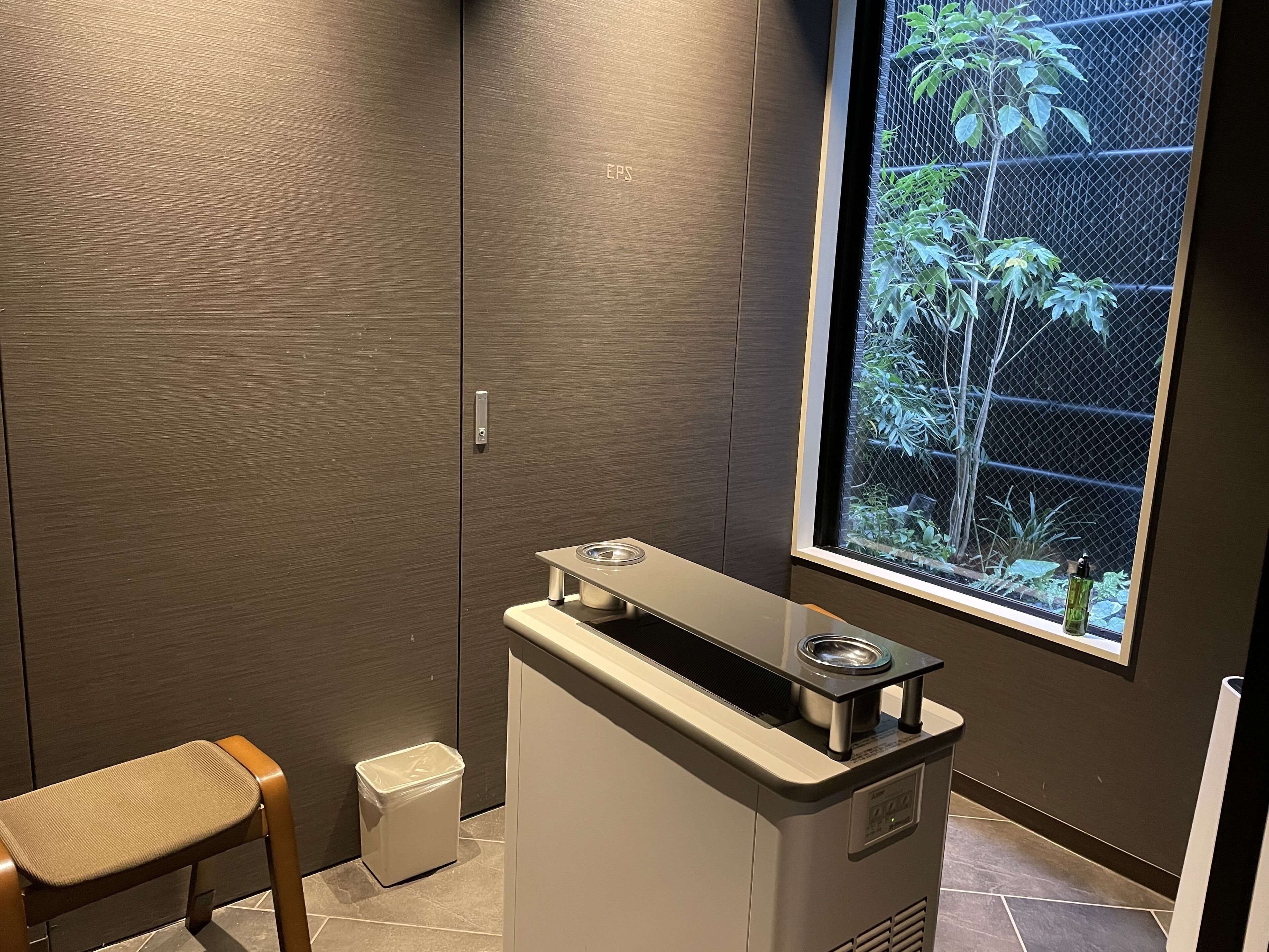 A bathroom with a wooden vanity, a sink with a mirror, a trash bin, and a window with a view of greenery.