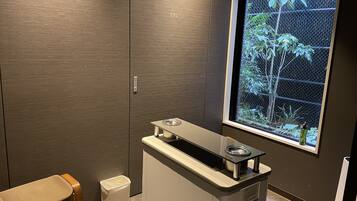 A bathroom with a wooden vanity, a sink with a mirror, a trash bin, and a window with a view of greenery.
