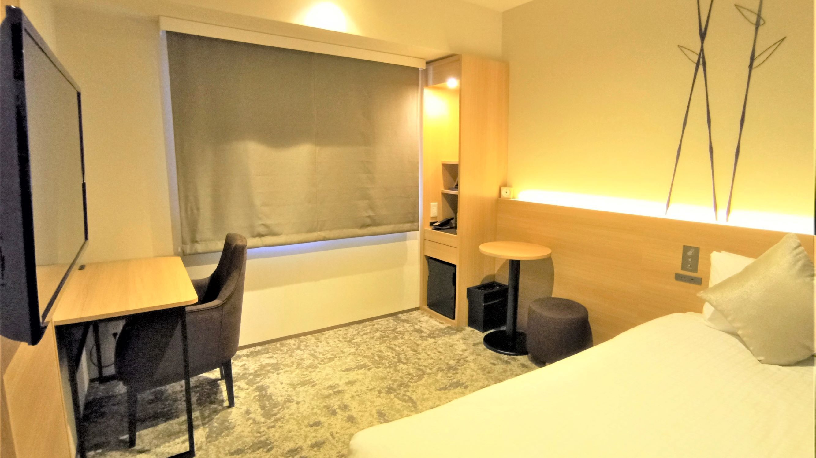 A hotel room with a bed, a desk, a chair, a small table, a trash bin, and a window with curtains.