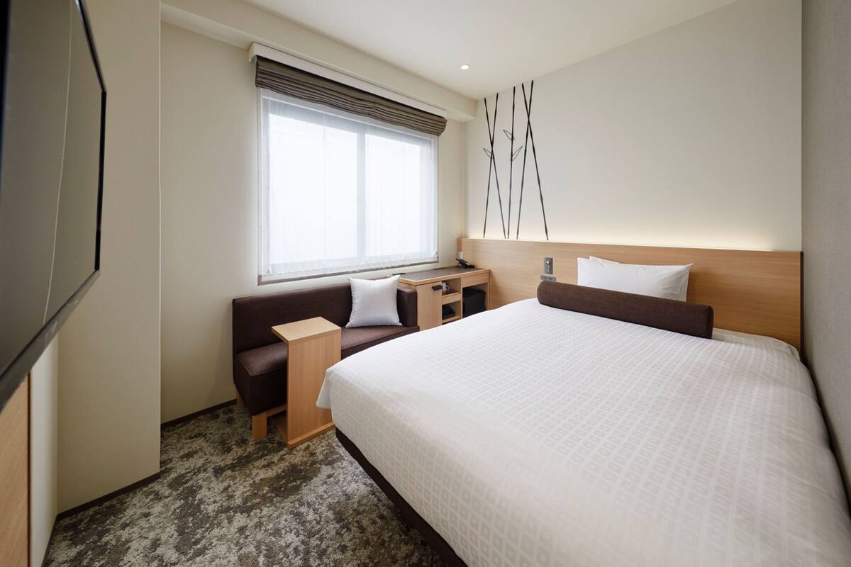 A hotel room with a large bed, a sofa, a desk, and a window with blinds.