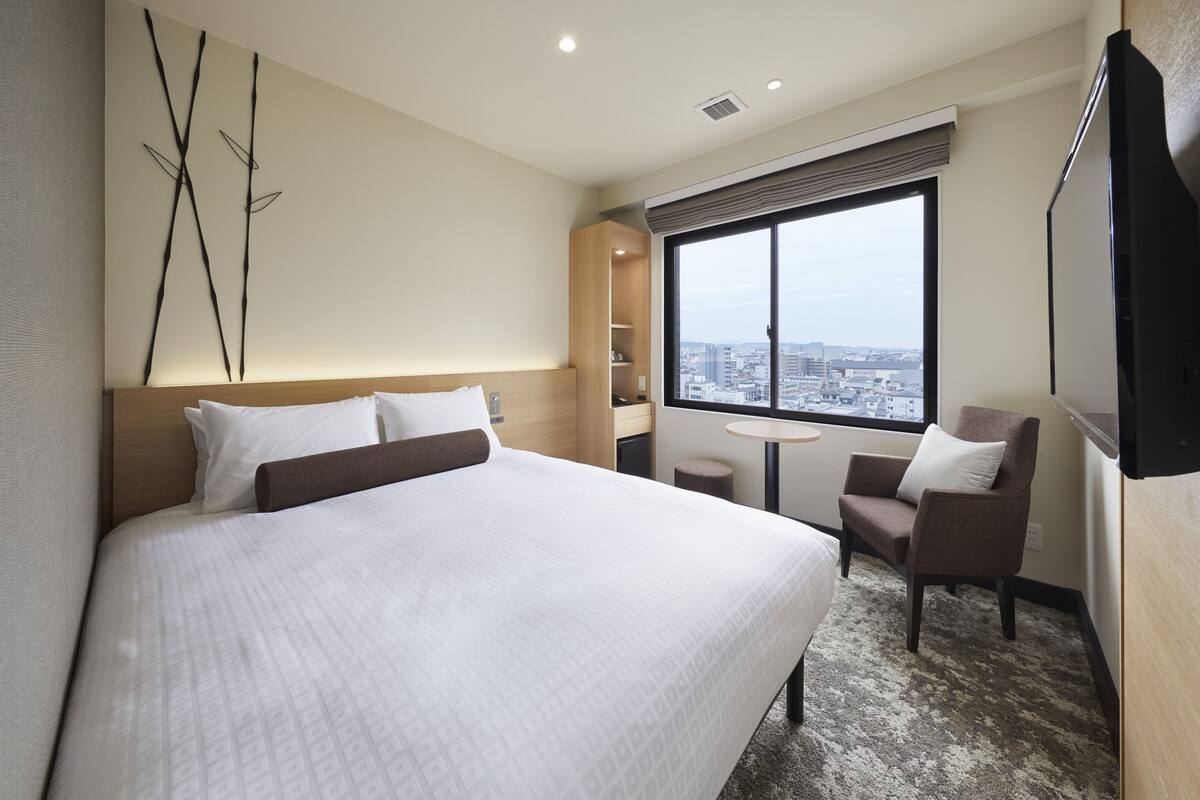A hotel room with a large bed, a small table, a chair, a TV, a window with a city view, and a bookshelf.