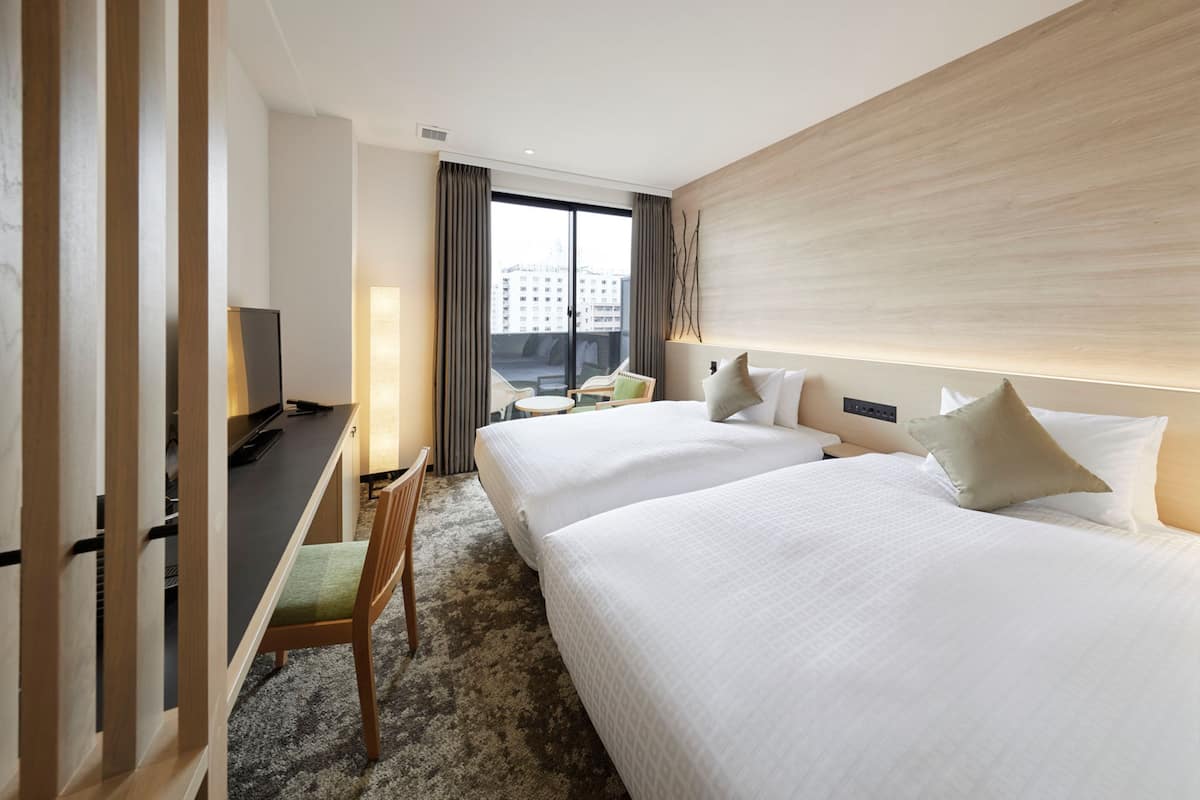 A hotel room with two beds, a desk with a chair, a TV, and a large window with a city view.
