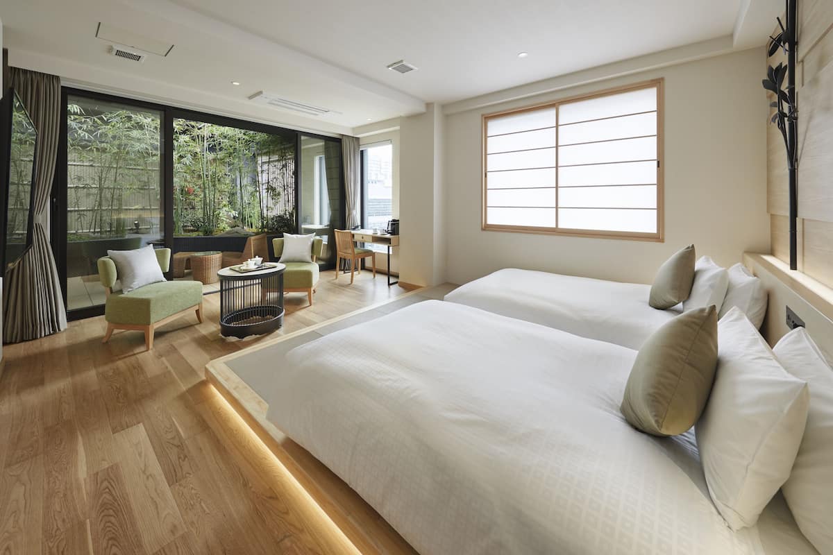 A modern hotel room with a large bed, a desk, and a view of greenery through large windows.