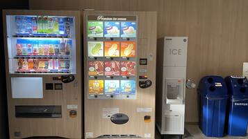 A vending machine with a variety of drinks and ice cream options, a small refrigerator labeled "ICE," and two blue trash bins.
