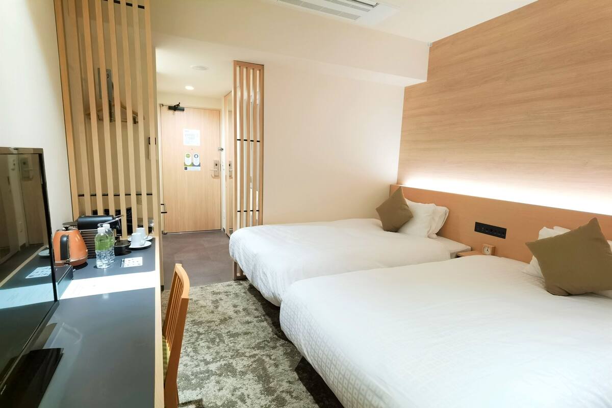 A hotel room with two beds, a desk with a kettle, a chair, and a wooden partition.