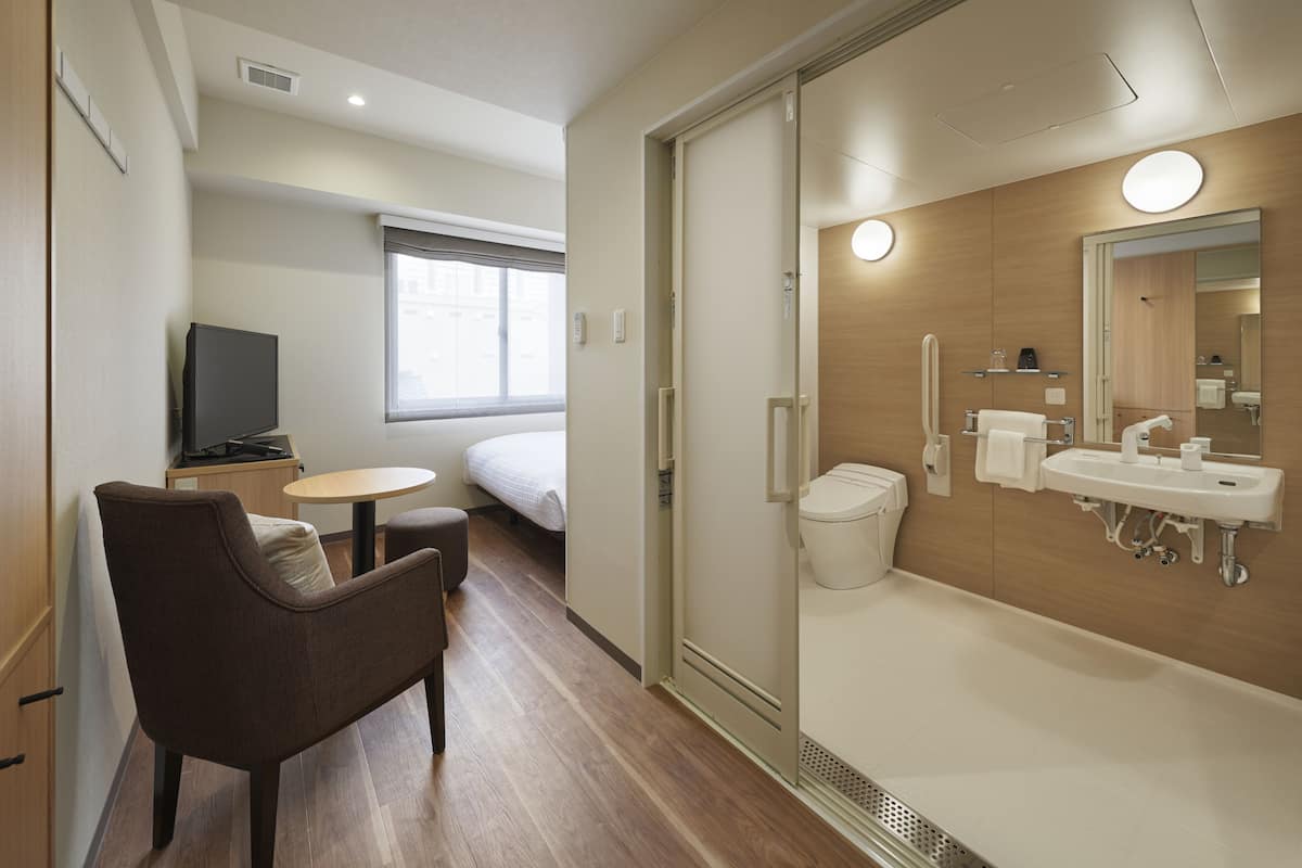 A modern hotel room with a bed, a small table with a chair, a TV, a shower area, and a bathroom with a sink and mirror.