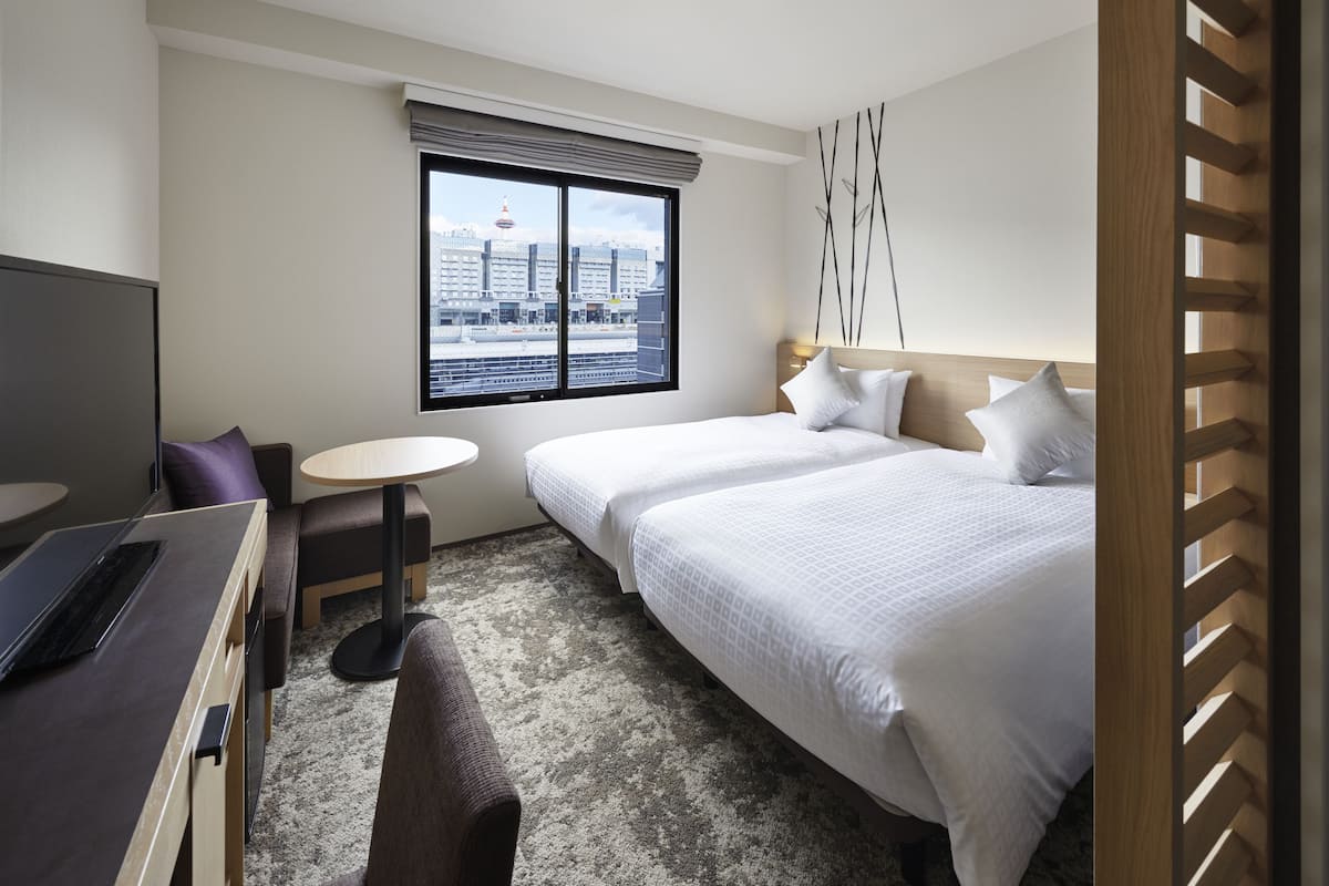A hotel room with two beds, a desk with a laptop, a small round table, and a view of the city through a large window.