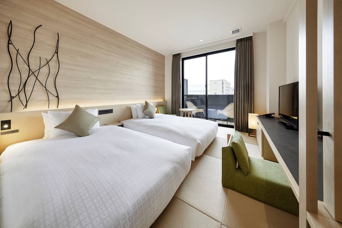 A modern hotel room with two large beds, a flat-screen TV, a small seating area, and a large window offering a city view.