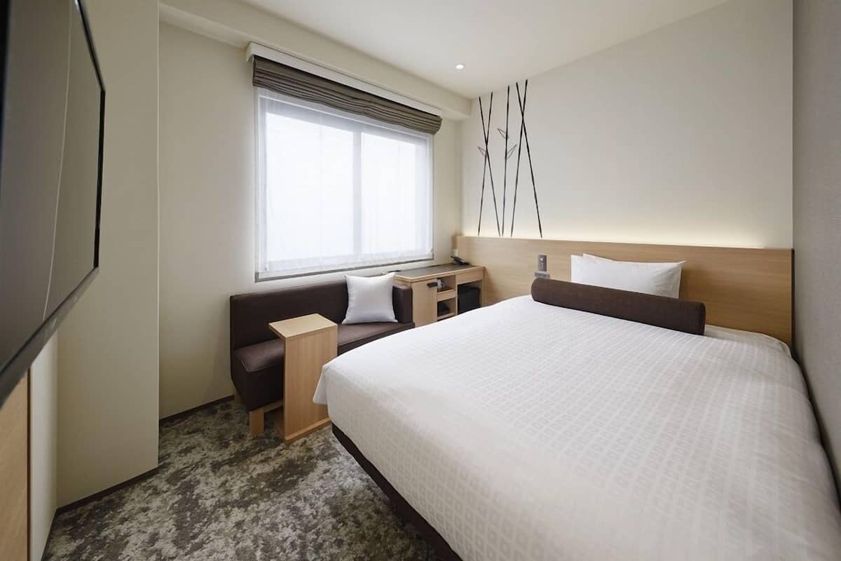 Hotellihuone, jossa on sänky, työpöytä, tuoli, sohva ja ikkuna, jossa on sälekaihtimet.