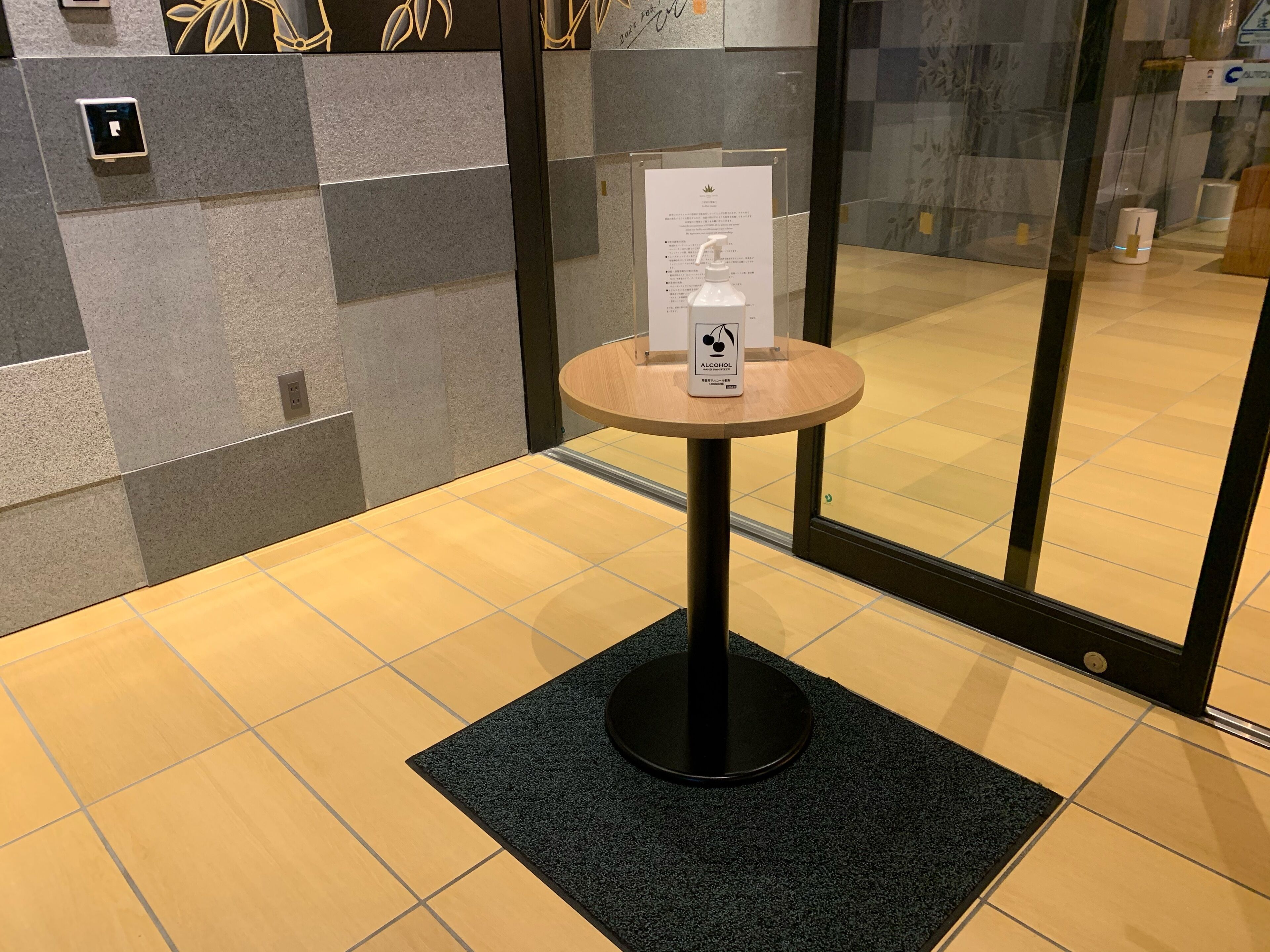 A reception area with a small round table displaying a bottle of alcohol and a sign, a tiled floor, and a glass door entrance.