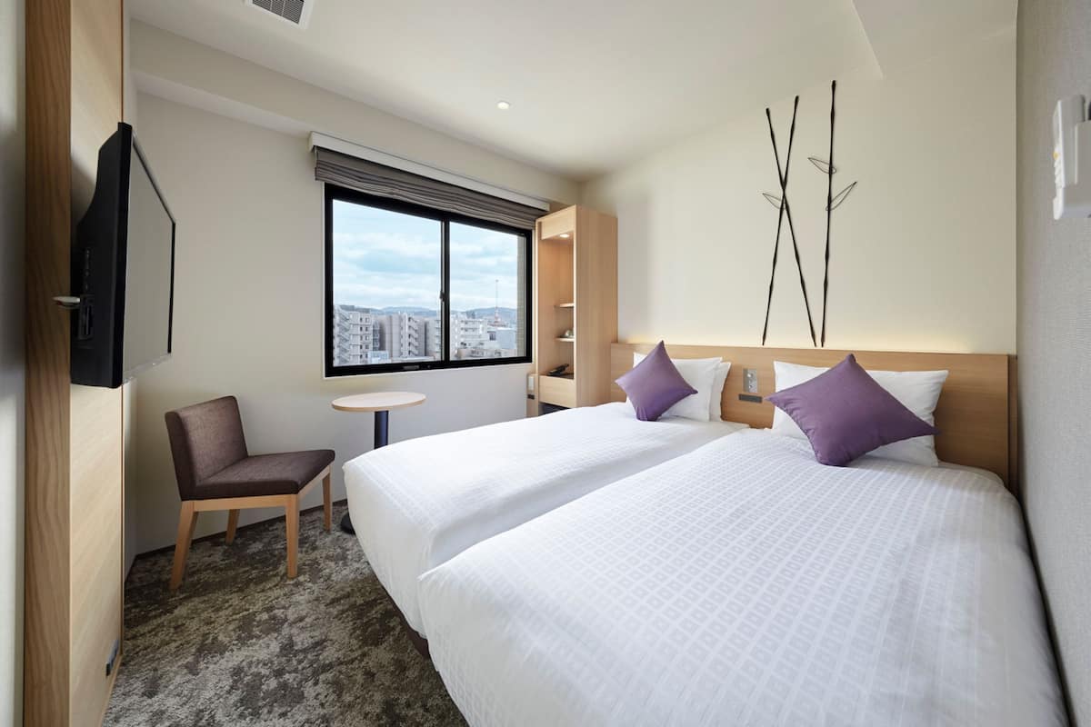 A hotel room with two beds, a desk, a chair, a TV, and a window with a city view.