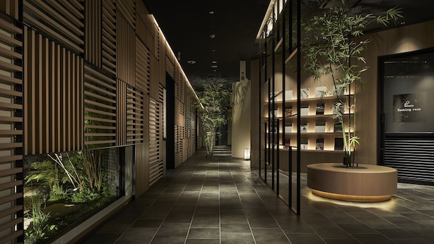 Un pasillo interior moderno con una recepción, plantas de bambú y una zona para fumadores.