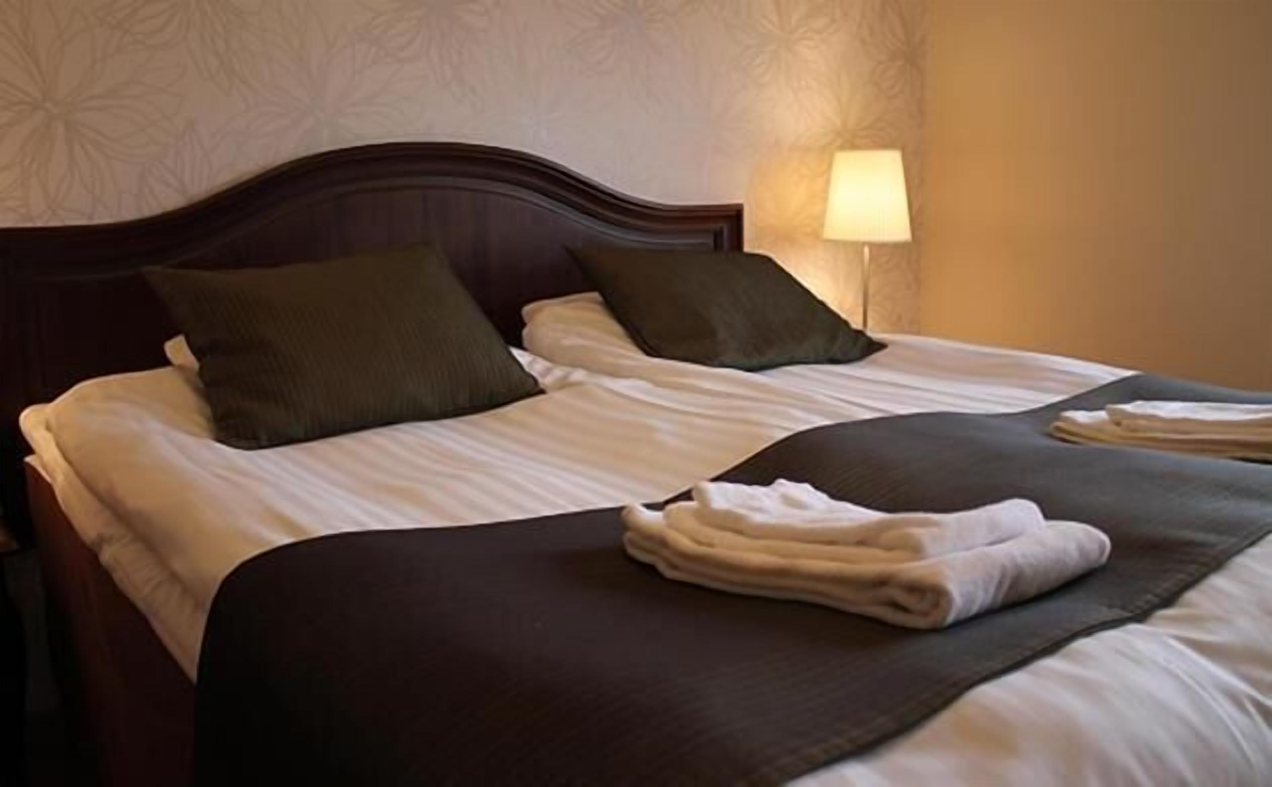 A neatly made bed with two pillows, a folded towel, and a bedside lamp.