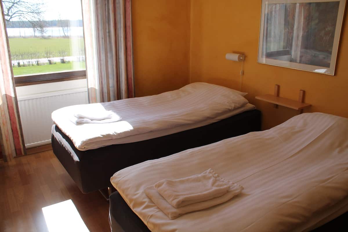 A hotel room with two single beds, a window with curtains, a painting on the wall, and a wooden floor.