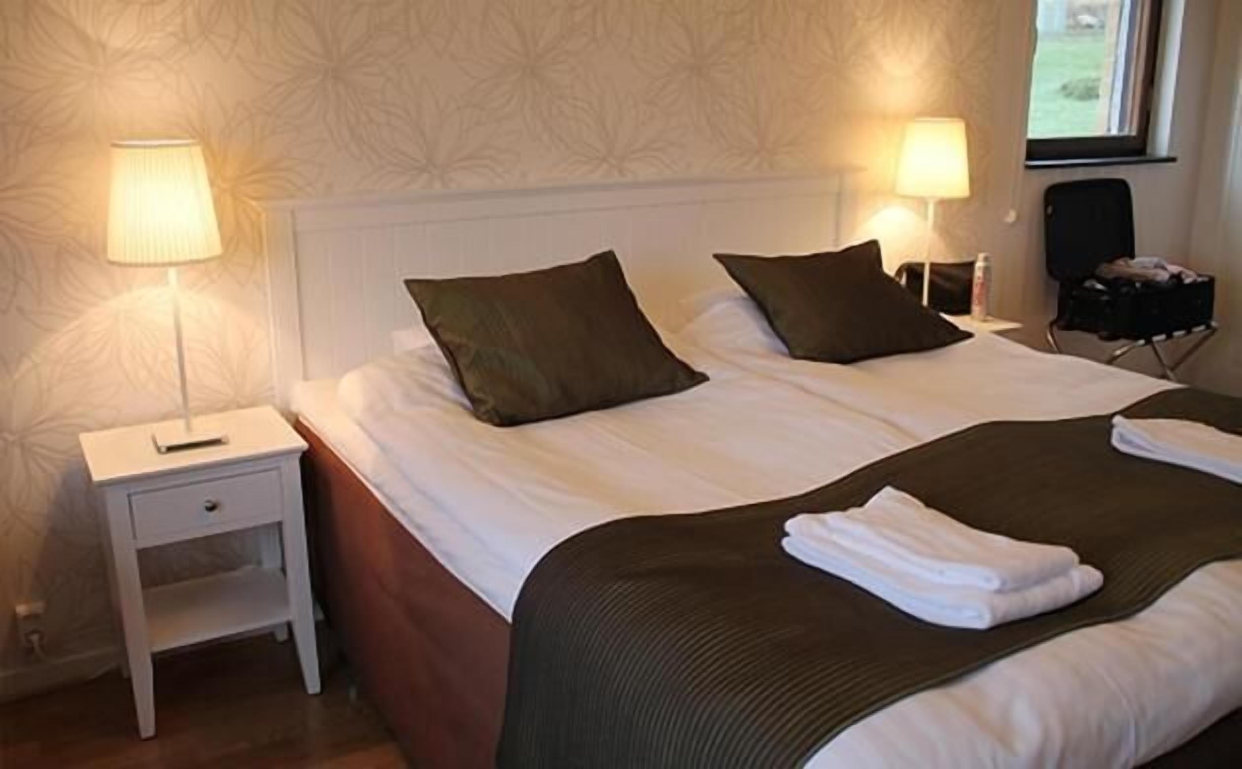 A neatly made bed with pillows, a bedside table with a lamp, and a suitcase on a chair.