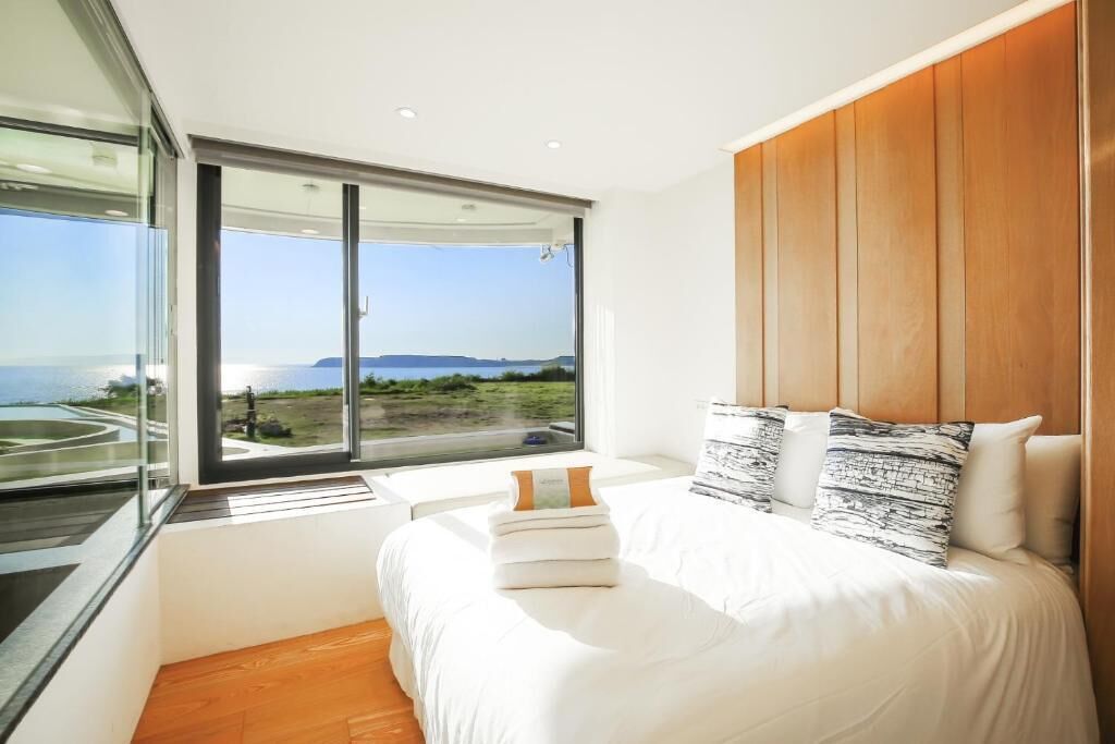 Un dormitorio moderno con una cama grande, cabecera de madera y vista al mar a través de ventanales de piso a techo.