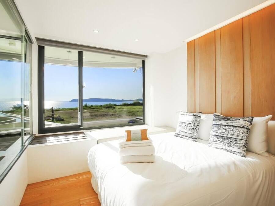 Un dormitorio moderno con una cama grande, cabecera de madera y vista al mar a través de ventanales de piso a techo.