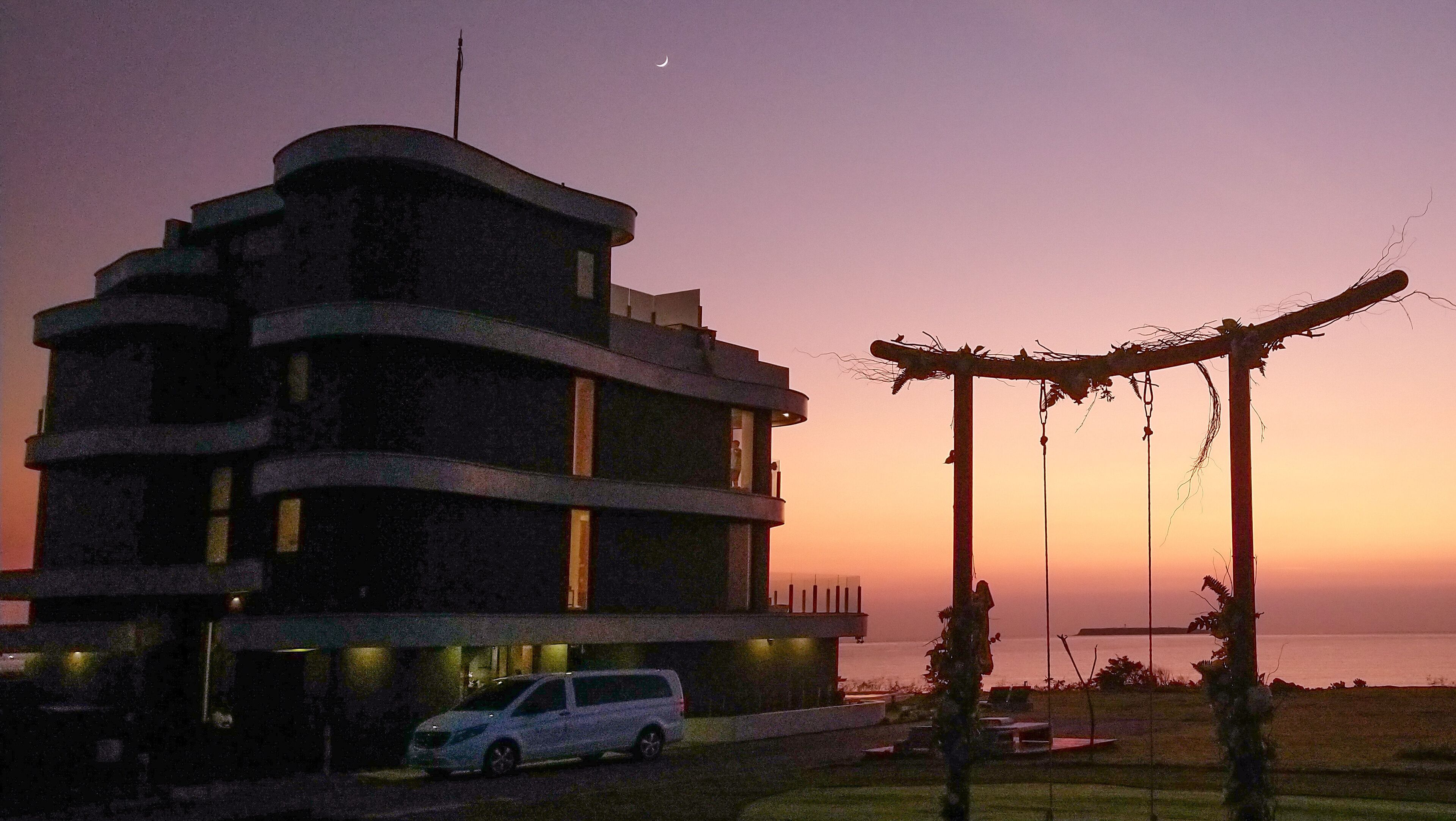 A modern building with a curved facade, a parked car, and a view of the sea at sunset.