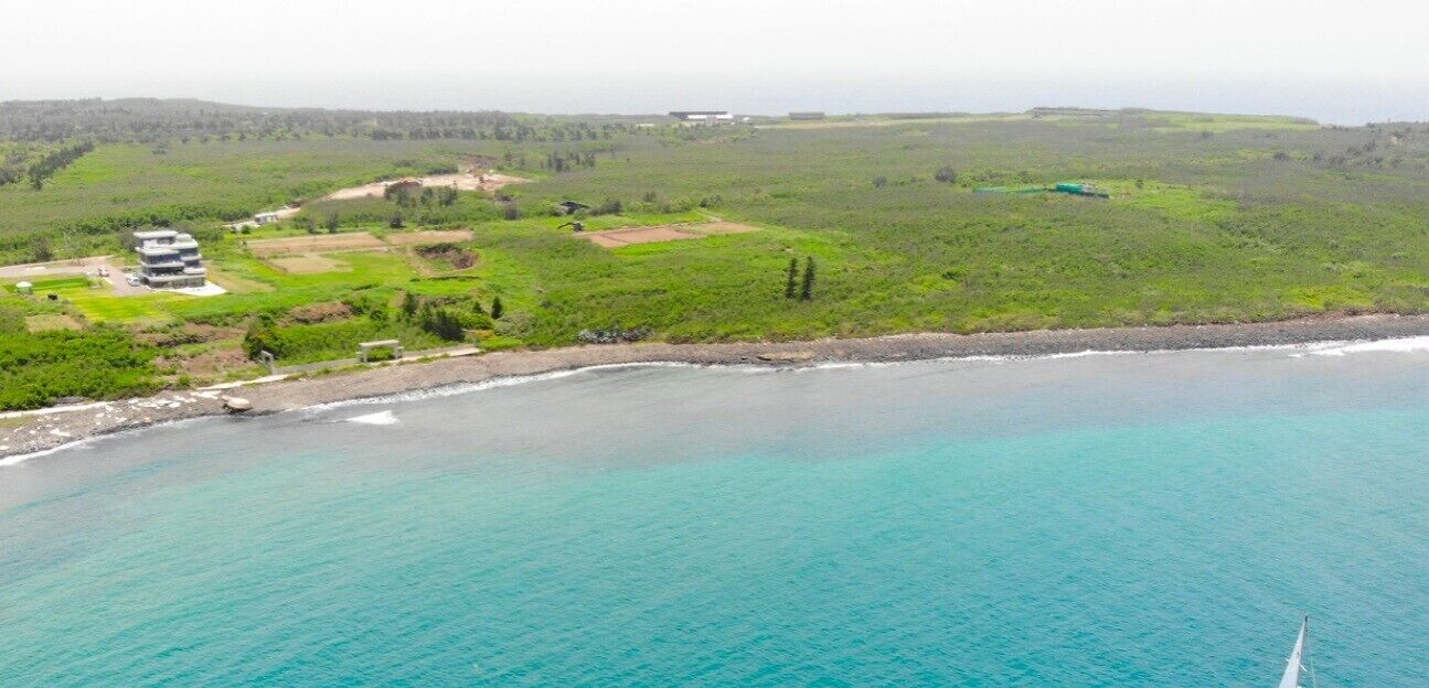 An aerial view of a coastal area with a building complex near the shore, surrounded by green fields and a clear blue sea.