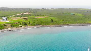An aerial view of a coastal area with a building complex near the shore, surrounded by green fields and a clear blue sea.