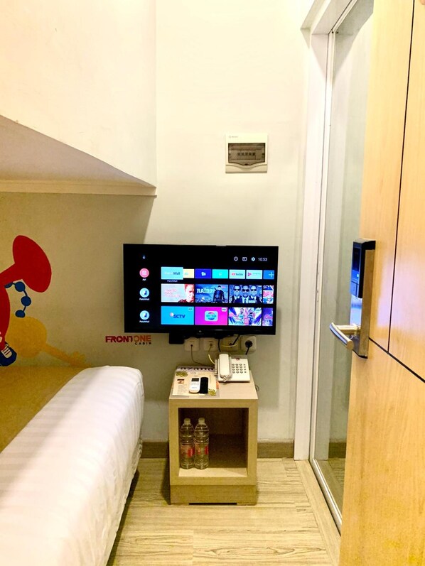 A hotel room with a bed, a TV mounted on the wall displaying various channels, a small table with a phone and a newspaper, and a wooden cabinet. - Front One Cabin Sunter (Jakarta)