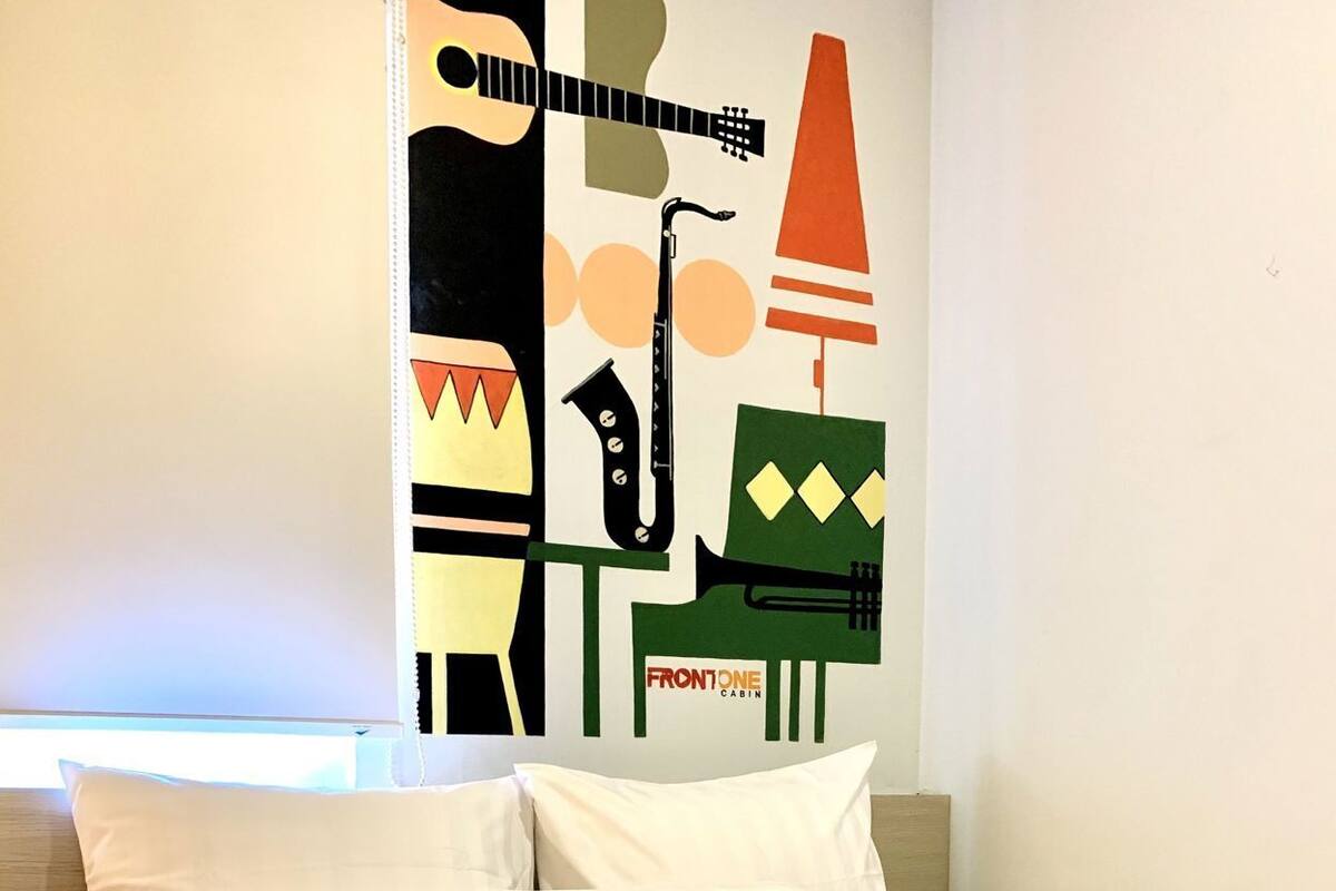 A bed with white bedding and pillows, a wall with a musical instrument mural, and a sign that reads 'FRONTONE CABIN'.