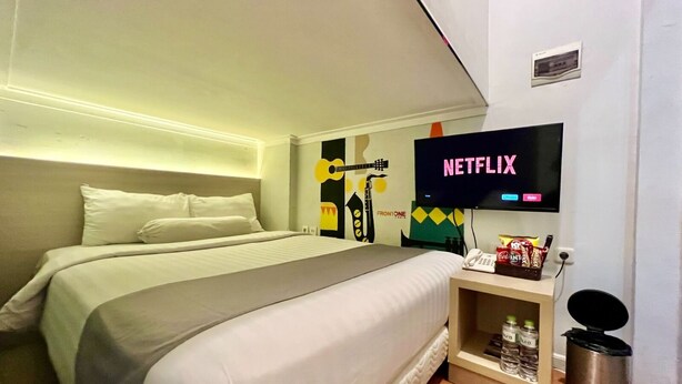 A hotel room with a bed, a television displaying 'NETFLIX', a small shelf with snacks, and a trash bin. - Front One Cabin Sunter (Jakarta)