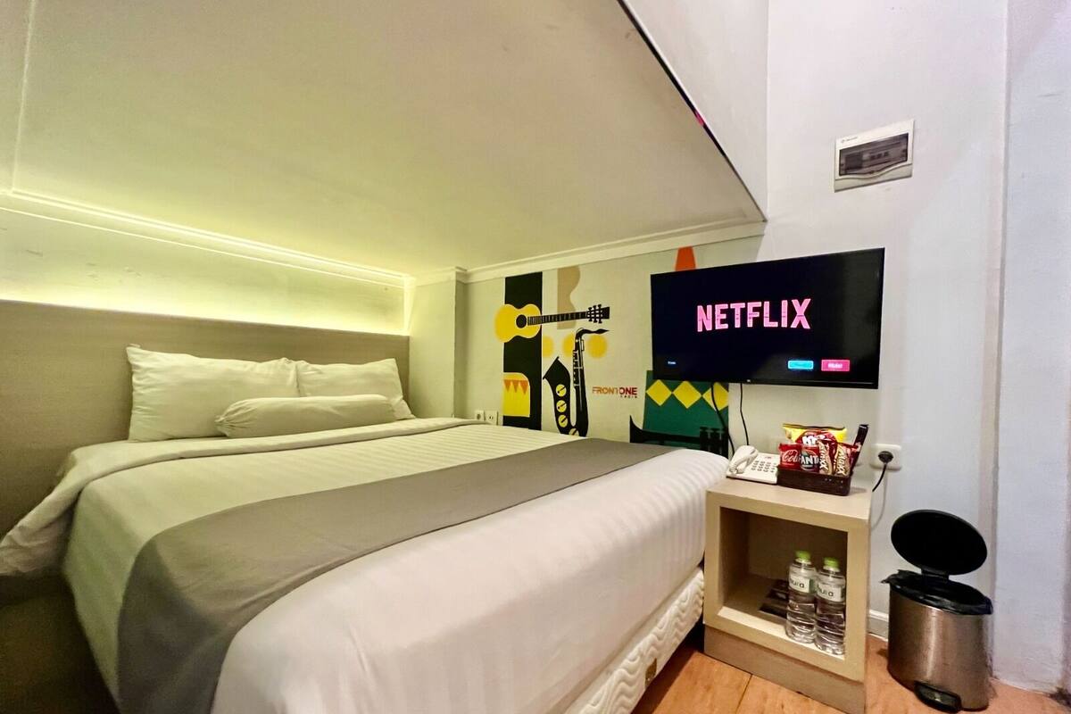 A hotel room with a bed, a television displaying 'NETFLIX', a small shelf with snacks, and a trash bin.
