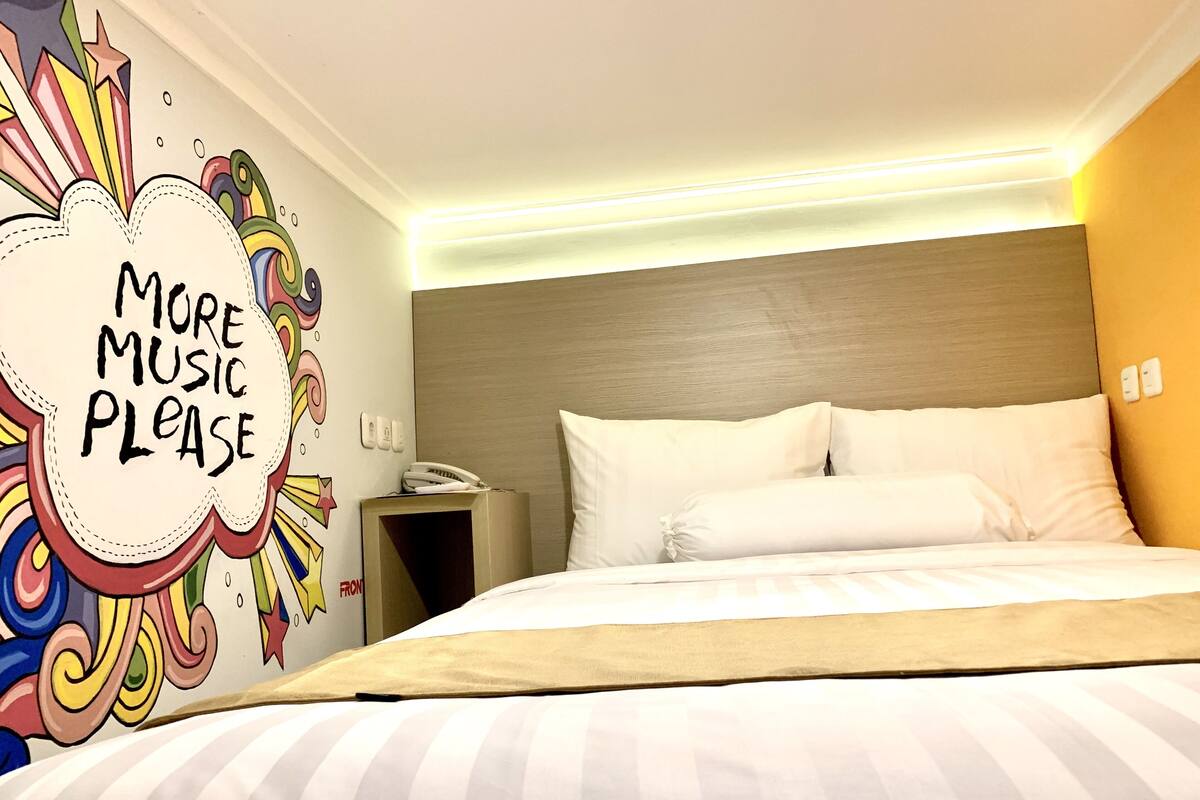 A hotel room with a bed, a bedside table with a phone, and a wall with a colorful mural saying "MORE MUSIC PLEASE".
