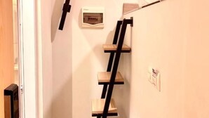 A narrow room with a wooden ladder shelf unit against the wall, a door, and a small table with bottles. - Front One Cabin Sunter (Jakarta)