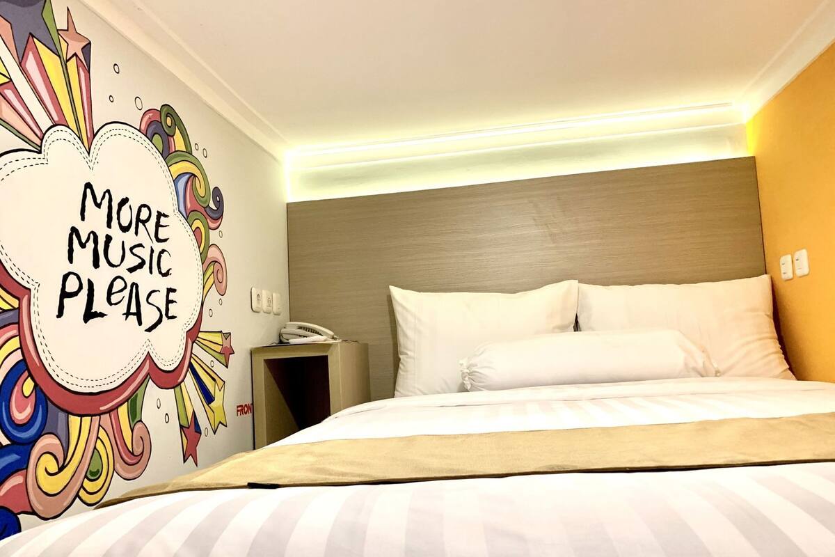 A hotel room with a bed, white pillows, a bedside table with a phone, and a wall with a colorful mural.
